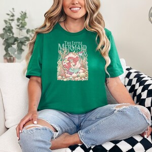 A woman wearing a green tee with "The Little Mermaid" text and an illustration of Ariel surrounded by underwater plants and sea creatures.