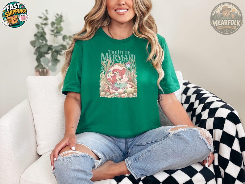 A woman wearing a green tee with "The Little Mermaid" text and an illustration of Ariel surrounded by underwater plants and sea creatures.