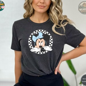 May include: Dark heather gray t-shirt featuring a Minnie Mouse graphic. The graphic shows Minnie Mouse with a light blue bow, set within a black and white checkered circle. The shirt is a crew neck style.