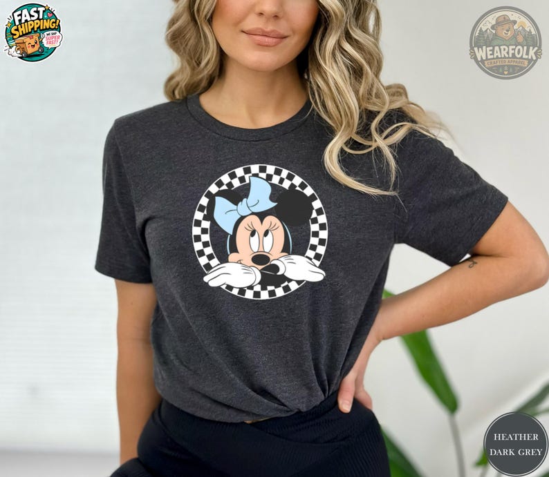 May include: Dark heather gray t-shirt featuring a Minnie Mouse graphic. The graphic shows Minnie Mouse with a light blue bow, set within a black and white checkered circle. The shirt is a crew neck style.