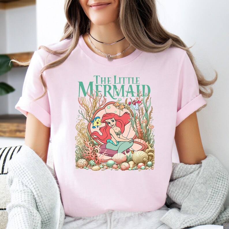 A woman wearing a light pink tee with "The Little Mermaid" text and an illustration of Ariel surrounded by underwater sea creatures and coral.
