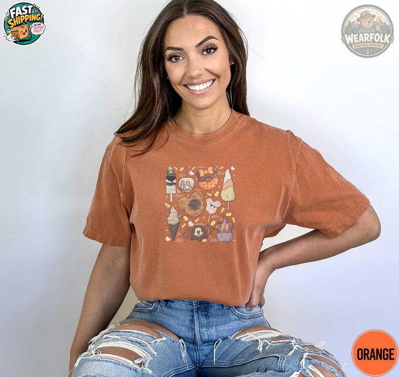 May include: Orange t-shirt featuring a square graphic with Halloween-themed treats, including ice cream cones, donuts, and candy corn. The shirt has short sleeves and a relaxed fit. The word "ORANGE" is printed on a circle at the bottom.