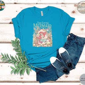 A woman wearing a turquoise tee with "The Little Mermaid" text and an illustration of Ariel surrounded by coral and sea creatures.