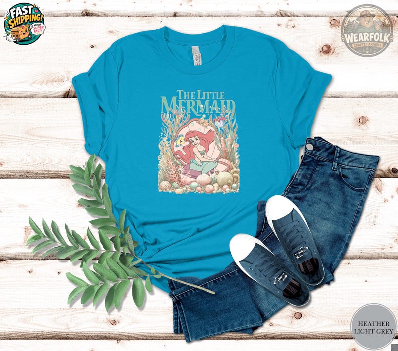 A woman wearing a turquoise tee with "The Little Mermaid" text and an illustration of Ariel surrounded by coral and sea creatures.