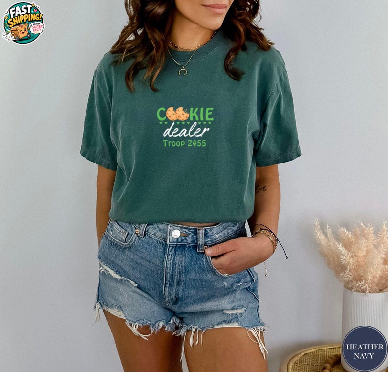 May include: A teal-colored t-shirt with the words "COOKIE dealer" in green and orange, and "Troop 2455" in green. The shirt is paired with denim shorts. The image also includes a small vase with dried flowers.