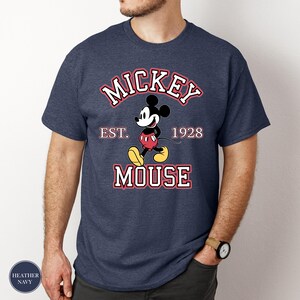 A man wearing a heather navy t-shirt featuring Mickey Mouse with bold varsity-style text reading "MICKEY EST. 1928 MOUSE" in white letters with red outlines.