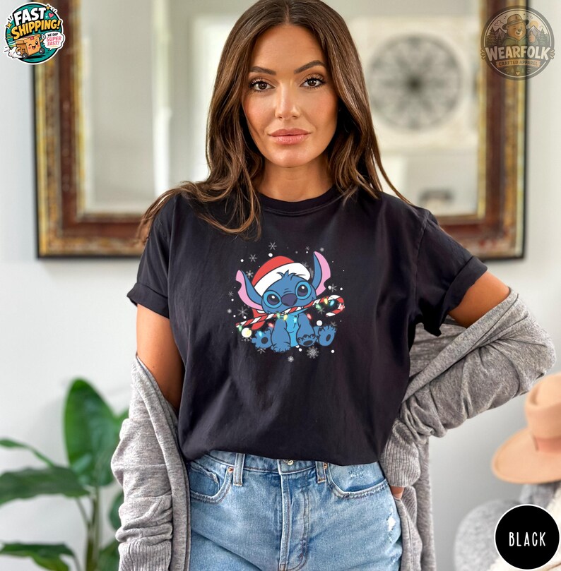 Woman wearing a black Bella Canvas tee with a blue alien character wearing a Santa hat holding a candy cane surrounded by snowflakes and ornaments.