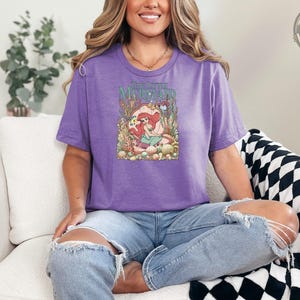 A woman wearing a purple tee with a colorful The Little Mermaid graphic featuring Ariel surrounded by underwater flowers and coral.