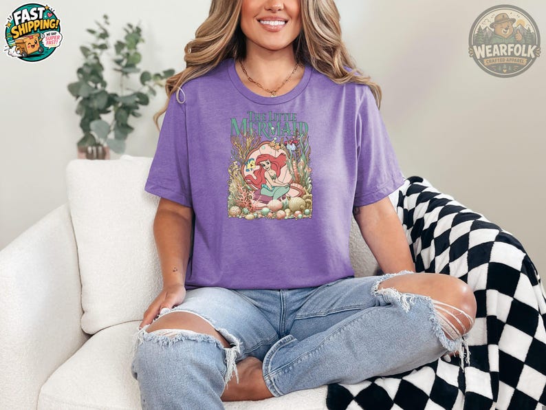 A woman wearing a purple tee with a colorful The Little Mermaid graphic featuring Ariel surrounded by underwater flowers and coral.