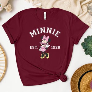 Flat lay of a heather maroon t-shirt featuring Minnie Mouse in sunglasses and polka dot dress, with bold white varsity text reading "MINNIE EST. 1928" on the chest.
