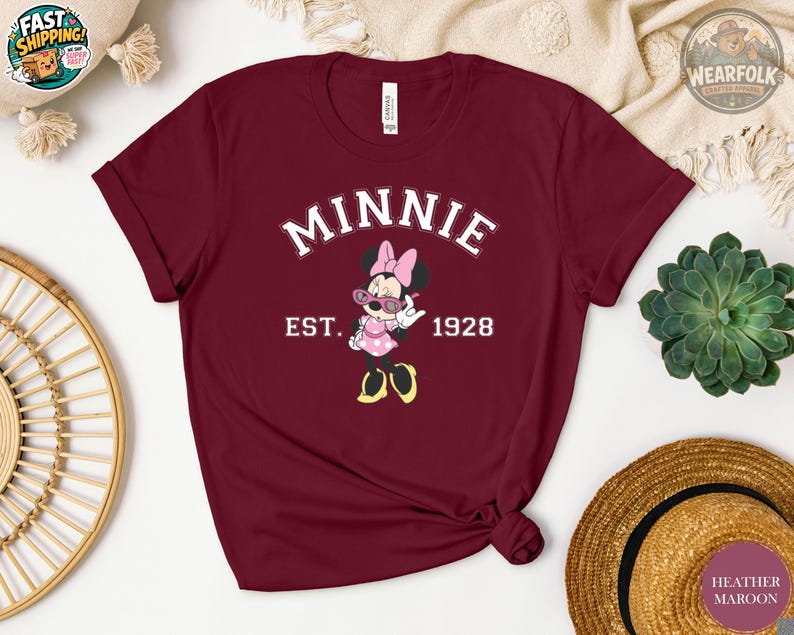 Flat lay of a heather maroon t-shirt featuring Minnie Mouse in sunglasses and polka dot dress, with bold white varsity text reading "MINNIE EST. 1928" on the chest.