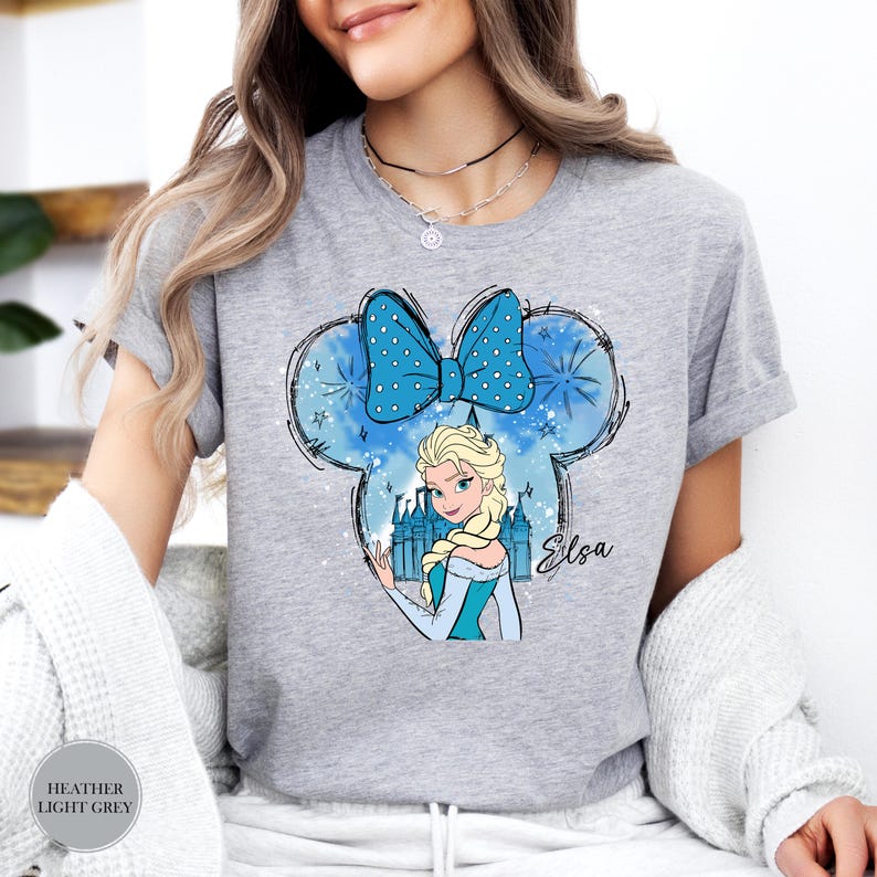 A woman wearing a heather grey tee with a blue Mickey Mouse head design featuring Elsa from Frozen, a castle, and the name "Elsa" in script.