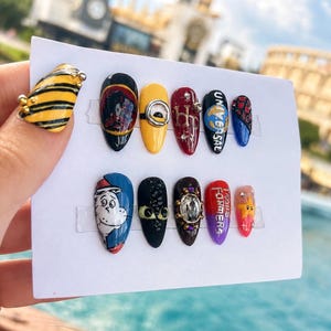 Puede incluir: Un juego de doce uñas postizas con varios diseños coloridos que incluyen un patrón de rayas amarillas y negras, un ojo de minion, el texto Universal, la marca Harry Potter, una ilustración del Gato en el Sombrero, una cara de dragón y el texto Transformers.