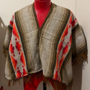 Könnte beinhalten: Ein gewebtes Poncho in Grau-, Braun- und Orangetönen, präsentiert auf einer roten Schaufensterpuppe. Der Poncho hat einen V-Ausschnitt, Streifenmuster und dekorative Fransen am unteren Rand. Das Design umfasst rote und orangefarbene geometrische Muster.