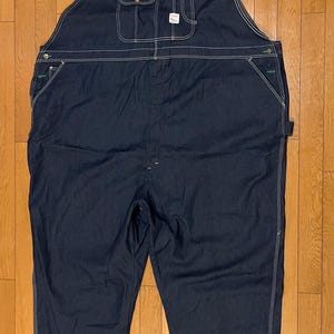 May include: Dark blue denim overalls with white stitching. Features include adjustable straps, multiple pockets, and a classic bib design. The overalls are made of durable fabric and are suitable for work or casual wear.