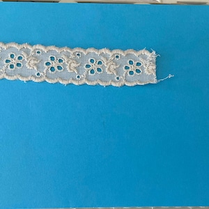 May include: Two pieces of white eyelet lace trim on a blue background. The lace trim has a scalloped edge and a floral design.
