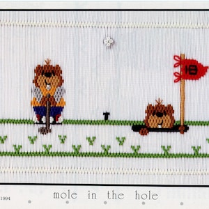 May include: A cross-stitch pattern featuring two brown moles playing golf. One mole is holding a golf club and the other is in a hole. The pattern includes green grass, a white golf ball, and a red flag with the number 18 on it. The text "mole in the hole" is printed below the pattern.