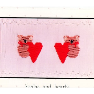 May include: A pink knitted baby blanket with two embroidered brown koalas holding red hearts. The text "koalas and hearts" is printed below the koalas.