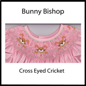 May include: Pink baby dress with a smocked neckline and embroidered bunnies. The dress has the text "Bunny Bishop" and "Cross Eyed Cricket" on it.