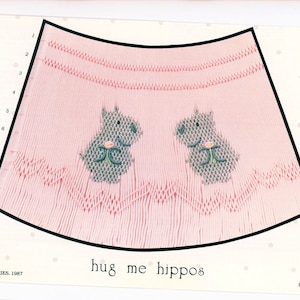 May include: A pink baby dress with a scalloped edge and two embroidered hippos. The dress has the text "hug me hippos" printed on the bottom.