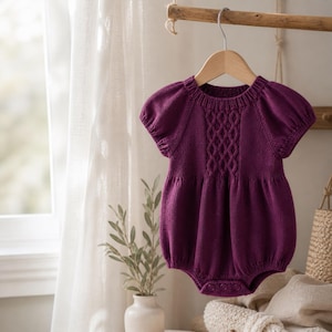 May include: A plum-coloured baby romper with short, puffed sleeves and a cable-knit design on the bodice. The knitted garment hangs from a wooden hanger, showcasing its soft texture and classic style. The romper is designed for comfort and style.