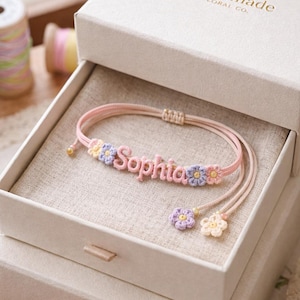 May include: A pink bracelet with the name "Sophia" in pink letters, adorned with small crochet flowers in pastel colors. The bracelet is presented in a white gift box with the text "Handmade Floral Co."