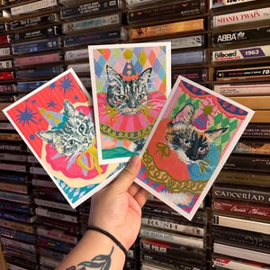 Könnte beinhalten: Drei farbenfrohe Katzenkunstdrucke. Jeder Druck zeigt eine Katze mit Partyhut und Rüschenkragen. Die Hintergründe sind lebendig mit geometrischen Mustern und Sternen. Die Drucke werden vor einer Wand aus Musikkassetten gehalten.