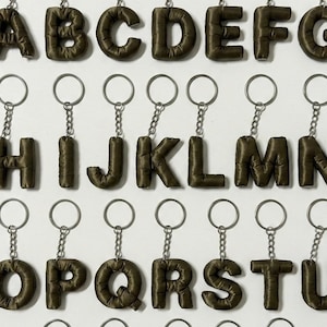 May include: Olive green fabric alphabet keychains, each letter attached to a silver keyring. The letters A through U are displayed, showcasing a unique, personalized accessory. The keychains are arranged against a white background.