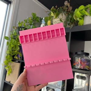 May include: A bright pink rectangular craft organizer with two rows of slots. The organizer is held in front of a window and shelves with plants and storage containers. The organizer is designed to hold small items.
