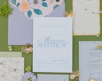 Op de afbeelding: Een platte presentatie van trouwbriefpapier op een groene achtergrond. Bevat een uitnodiging, een RSVP-kaart en een detailkaart. Een blauwe fluwelen ringdoos, een parelketting en een Gucci Flora parfumfles zijn ook aanwezig. Bloemenaccenten en goudkleurige decoratieve elementen maken de opstelling compleet.