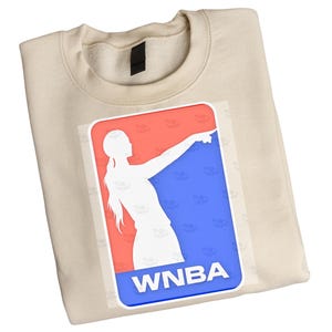 Op de afbeelding: Een beige sweater met ronde hals en een rechthoekige afbeelding op de borst. De afbeelding toont een wit silhouet van een basketballer die wijst, tegen een rode en blauwe achtergrond met de witte tekst WNBA onderaan gedrukt.