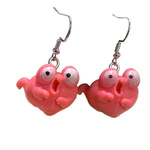 May include: A pair of dangle earrings featuring pink, cartoon-style creatures with large, wide-set white eyes and black pupils. The creatures have small arms, open mouths, and are suspended from silver-toned metal fishhook-style earring wires.