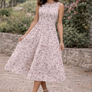 Puede incluir: Vestido sin mangas con estampado floral en tonos rosa y blanco. El vestido tiene un corpiño ajustado y una falda fluida, y se combina con sandalias de tiras blancas. El fondo presenta vegetación y un muro de piedra.