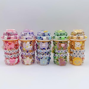 May include: Five decorative pill bottle containers, each adorned with colorful beads and charms. The bottles feature various designs, including an elephant, a bunny, a bear, and the words "LITTLE HELPER", "THANKS", "CHILL PILLS", "FOCUS", and "BADDIES NOT SADDIES".