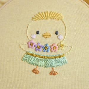 Puede incluir: Arte de aro de pollito bordado. El pollito es amarillo con un peinado de flecos amarillos, ojos negros y un pico amarillo. Lleva un collar de flores y una falda verde. El bordado está sobre tela amarilla en un aro de madera.