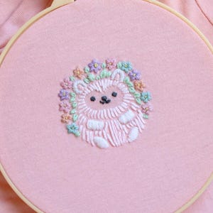 Może przedstawiać: Różowy tamborek do haftu z ręcznie wyszytym jeżem. Jeż jest biały z czarną twarzą i nosi wianek z kwiatów w pastelowych kolorach. Tło to miękka różowa tkanina, widoczna jest drewniana rama tamborka.