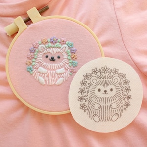 May include: A pink embroidery hoop with a hedgehog design, surrounded by a floral wreath. The hedgehog is white with a brown face and is surrounded by a wreath of colorful flowers. A matching hedgehog design is printed on a round, off-white fabric.