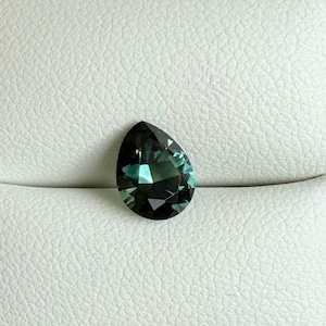 May include: A pear-shaped gemstone with a dark teal hue, displaying facets that catch the light. The gem has a polished surface and is set against a white background. The overall appearance is elegant and refined.