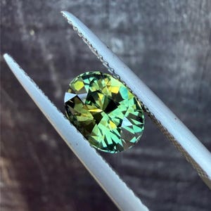 Puede incluir: Una gema de corte ovalado con una vibrante mezcla de tonos verdes y amarillos, sujeta por unas pinzas plateadas. Las facetas de la gema reflejan la luz, creando un efecto brillante. El fondo es borroso y de tono neutro.