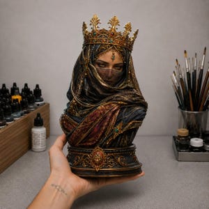Puede incluir: Un busto de resina detallado de una figura con una corona dorada ornamentada con joyas rojas y verdes. La figura está envuelta en un tocado oscuro y texturizado y un velo que oculta la parte inferior del rostro, sobre una base circular decorada.
