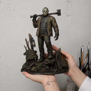May include: A detailed figurine of a character wearing a hockey mask, a brown jacket, and dark pants, holding an axe over one shoulder. The base features a sign that reads "WELCOME TO CAMP CRYSTAL LAKE," a hanging lantern, a skull, and tree roots.