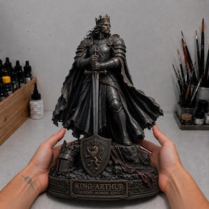 May include: A detailed dark bronze-toned statue of King Arthur wearing a crown and plate armor, holding a long sword. A shield with a lion emblem and a helmet sit at the base. A plaque reads: KING ARTHUR LEGEND. HONOR. KING.