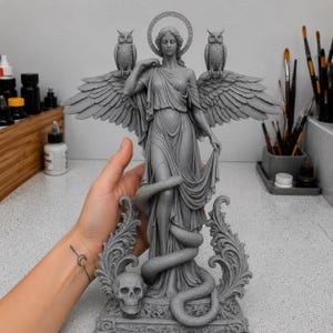 May include: A gray resin statue of a winged figure with a halo, standing with a snake coiled around the legs. The figure is flanked by two owls perched on the wings, with a skull at the base. The sculpture features intricate drapery and scrollwork.