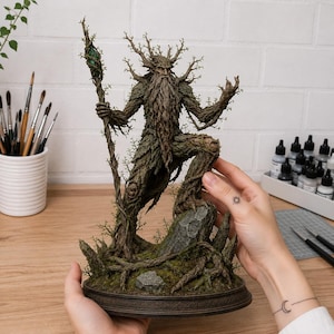 May include: A detailed figurine of a forest guardian creature with a bark-like texture, long beard, and branch-like antlers. The figure stands on a mossy base, holding a tall staff topped with a green crystal, rendered in shades of brown and green.