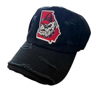Puede incluir: Gorra de béisbol negra desgastada con un parche rojo y blanco. El parche presenta un gráfico de bulldog y la letra "G" dentro de un contorno rojo del estado de Georgia. La gorra tiene una visera curva y un aspecto desgastado.
