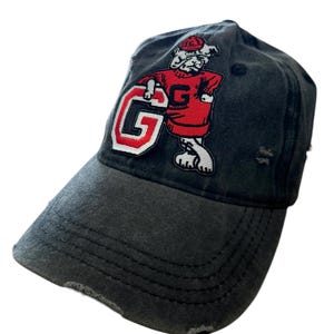 Puede incluir: Gorra de béisbol gris carbón desgastada con un parche rojo y blanco con una mascota de bulldog y la letra "G". La gorra tiene una visera curva y un aspecto desgastado, con detalles desgastados.
