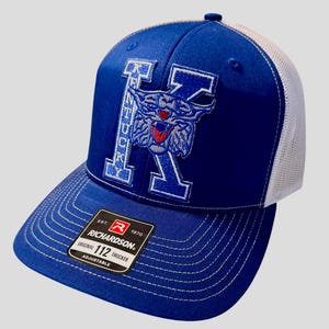 May include: A blue and white trucker cap featuring the letter "K" and a wildcat emblem. The cap has a curved peak and a white mesh back. The Richardson brand label is visible.