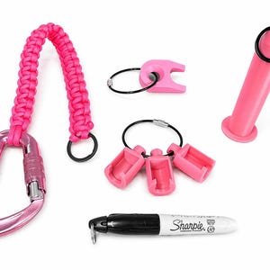 Puede incluir: Una colecci&oacute;n de accesorios rosas, que incluye un mosquet&oacute;n con asa trenzada, llaveros y un rotulador Sharpie. El mosquet&oacute;n tiene el texto "KN-1213-3". El Sharpie es negro con tapa blanca y el logotipo de Sharpie.