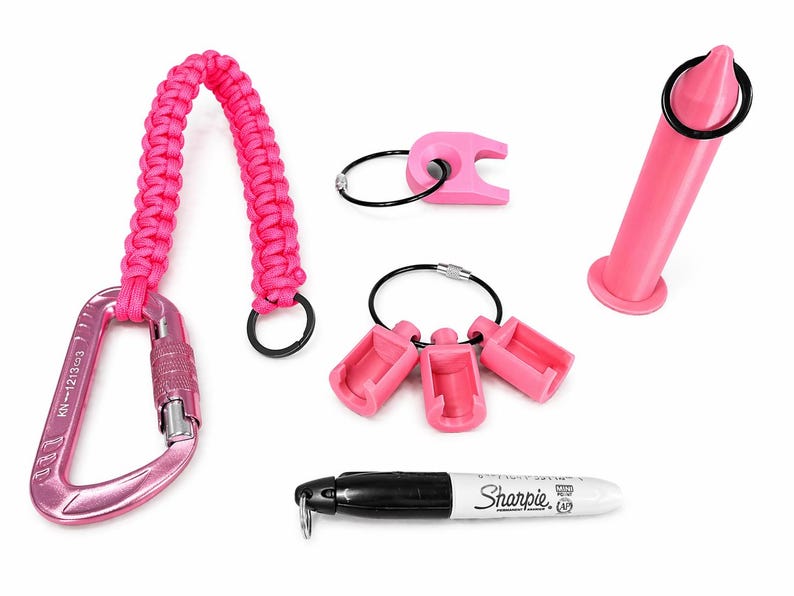Puede incluir: Una colecci&oacute;n de accesorios rosas, que incluye un mosquet&oacute;n con asa trenzada, llaveros y un rotulador Sharpie. El mosquet&oacute;n tiene el texto "KN-1213-3". El Sharpie es negro con tapa blanca y el logotipo de Sharpie.