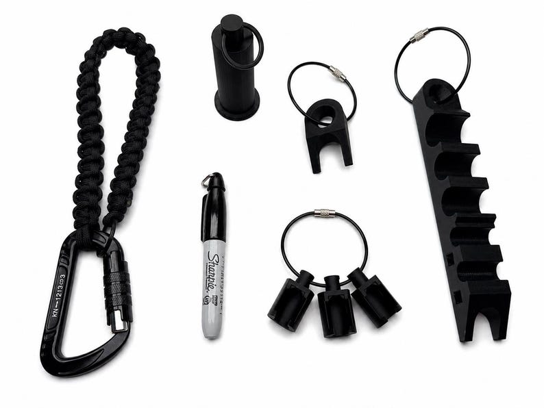 May include: A collection of black tactical gear including a carabiner with a braided paracord lanyard, a Sharpie marker, a cylindrical tool, a single-slot holder, a three-slot holder, and a long multi-slot organizer, all featuring metal rings.